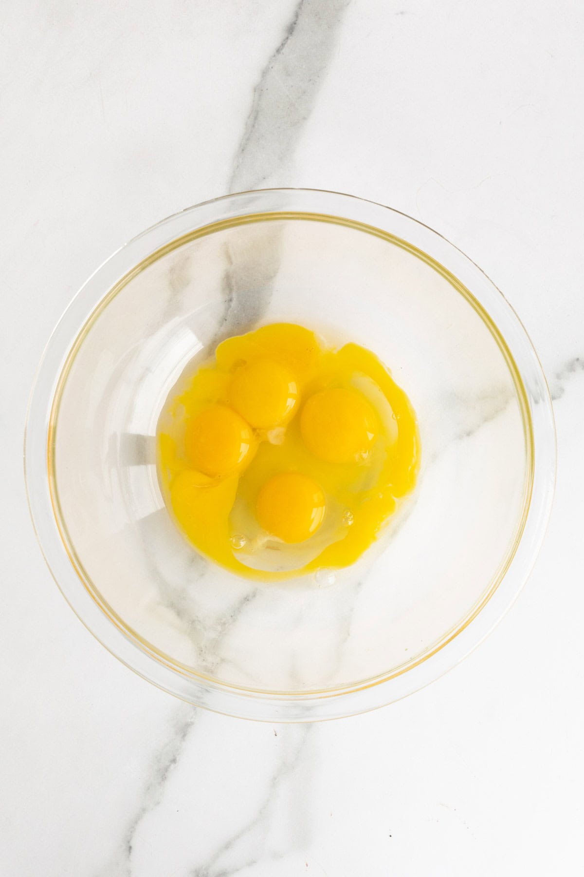 Four raw egg yolks in a clear glass bowl on a marble surface