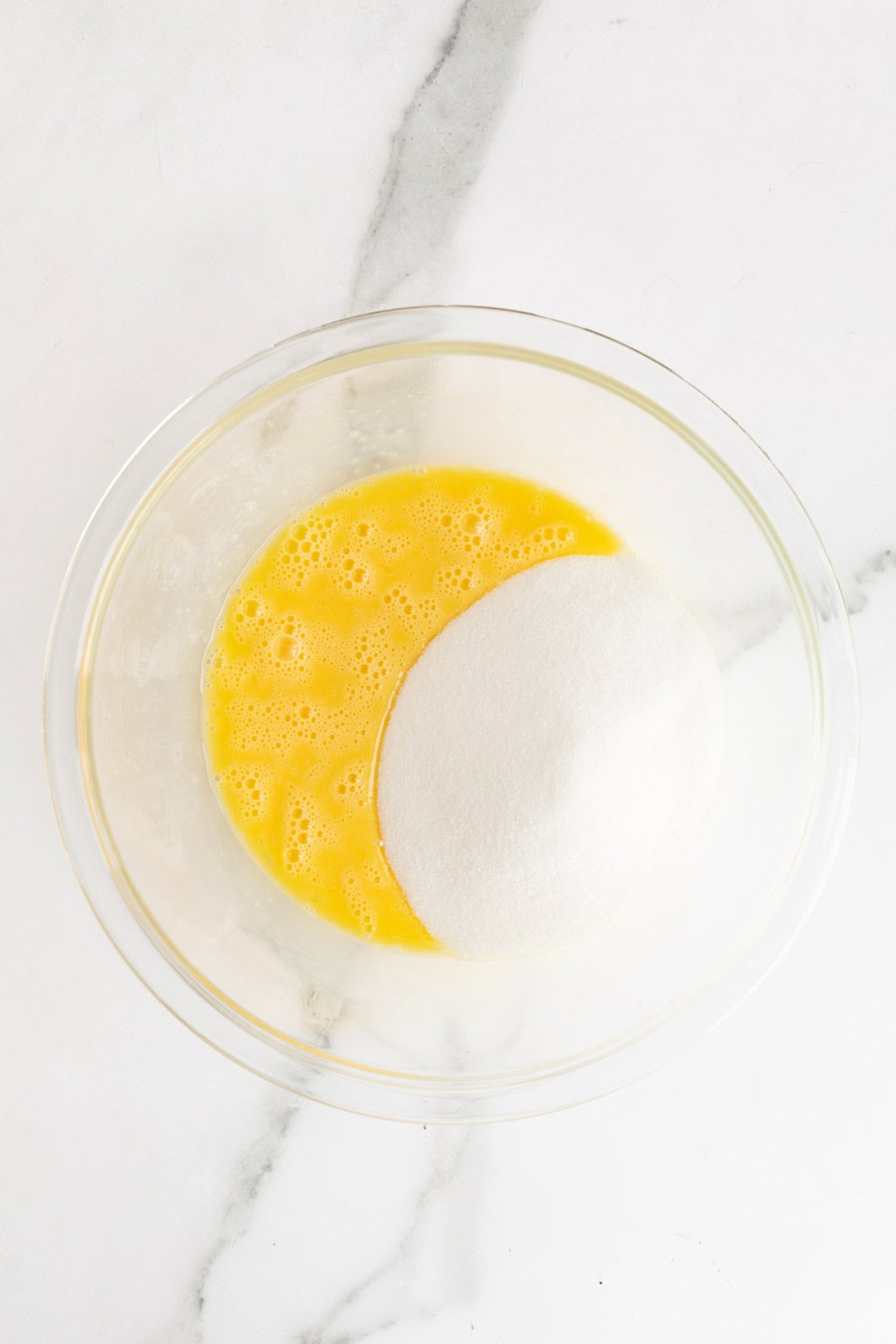 Granulated sugar added to whisked eggs in a clear bowl, partially mixed
