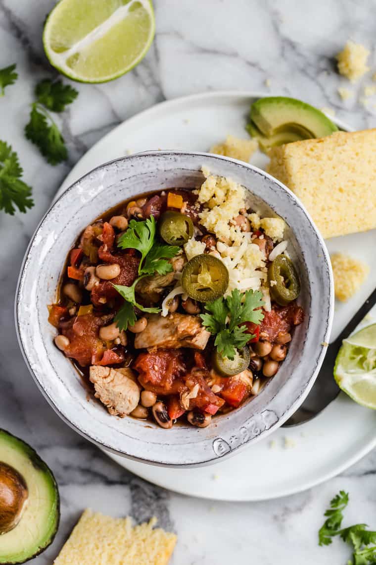 Overhead shot of a bowl of Chicken Chili with Black Eyed Peas with a slice avocado, lime and cornbread surrounding it