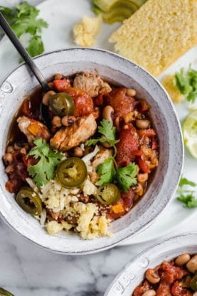 Overhead shot of a close up of a bowl of Chicken Chili with Black Eyed Peas with a slice avocado, lime and cornbread surrounding it