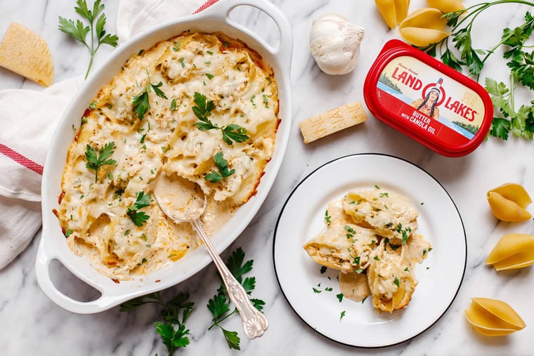 Overhead shot of chicken stuffed shells recipe served in a white casserole dish with a spoon and an individual serving served on a white dish next to it along with uncooked pasta shells and a container of Land O Lakes butter