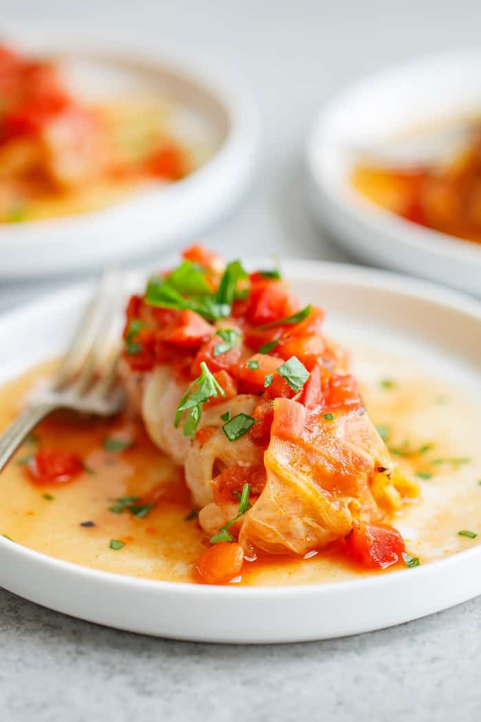 Stuffed cabbage roll recipe with tomato sauce and parsley garnish on white plate with fork ready to serve with other plates in background