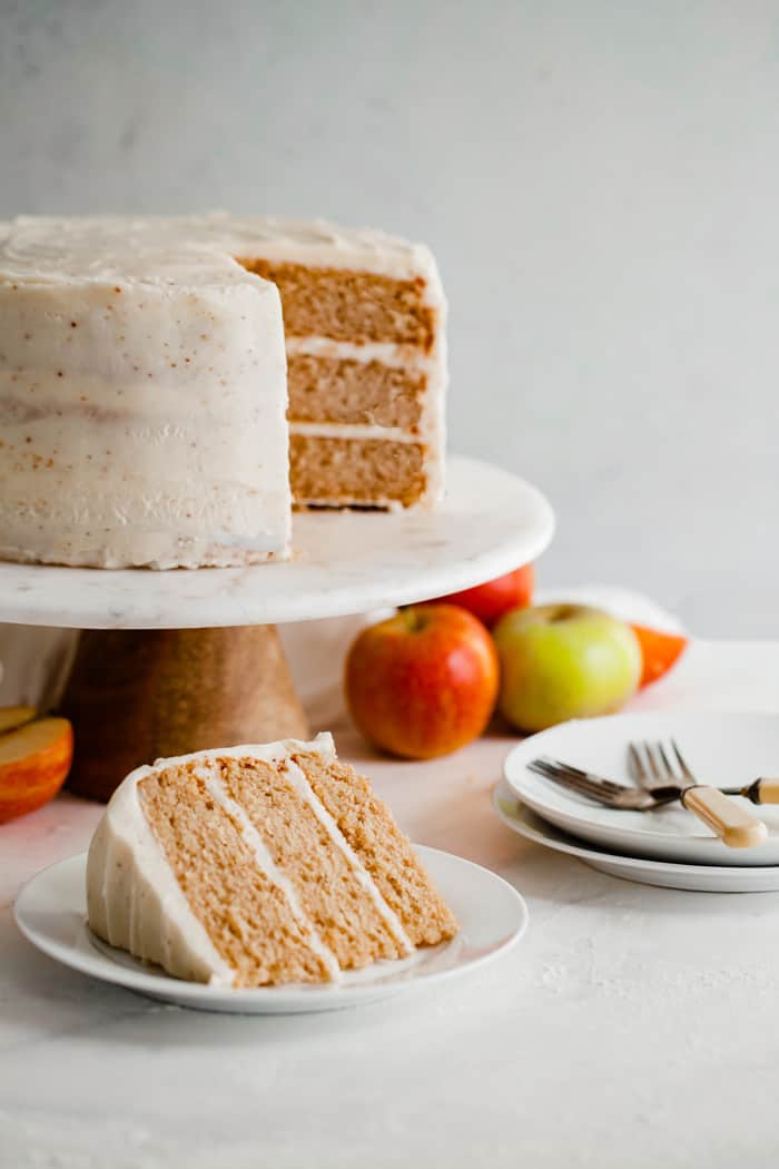 Spiced Cider Apple Cake with Brown Butter Frosting sliced and displayed on a cake plate with one slice served on a white plate with apples surrounding