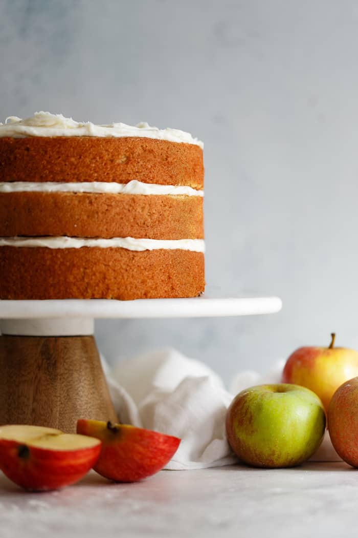 Three layer Spiced apple cider Cake with Brown Butter Frosting displayed on a cake plate with apples in the foreground
