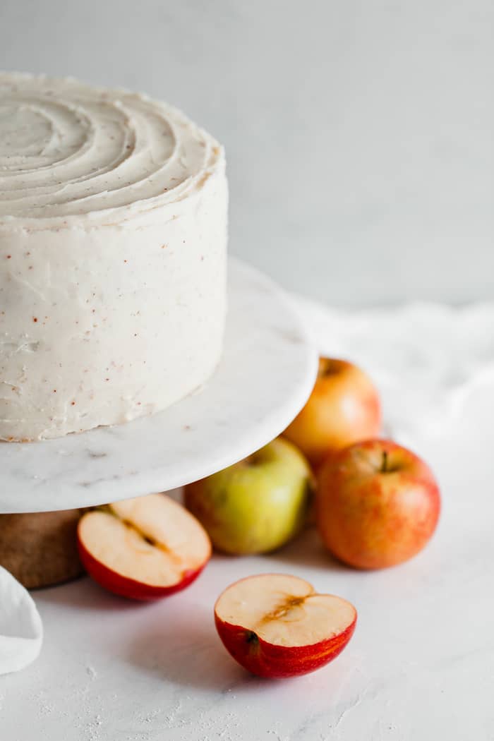 Spiced cider apple cake recipe iced in brown butter frosting on a white cake stand with apples surrounding it