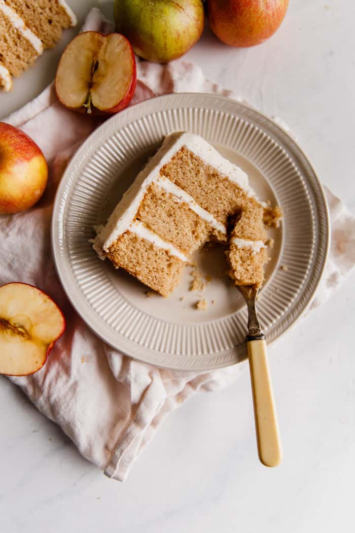Overhead shot of a slice of Spiced Apple Cider Cake with Brown Butter Frosting served on a circular plate with a fork and sliced apples next to it