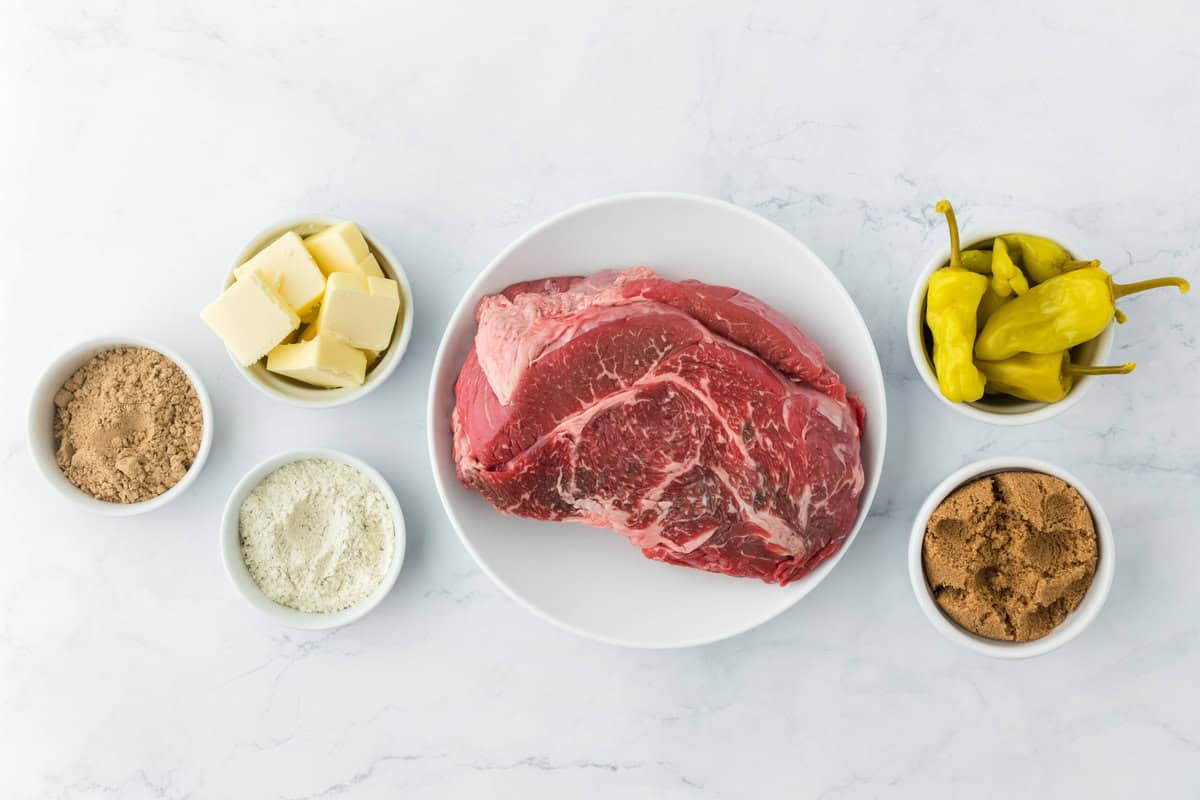 A chuck roast along with other ingredients to make a Mississippi roast on a white background in white bowls