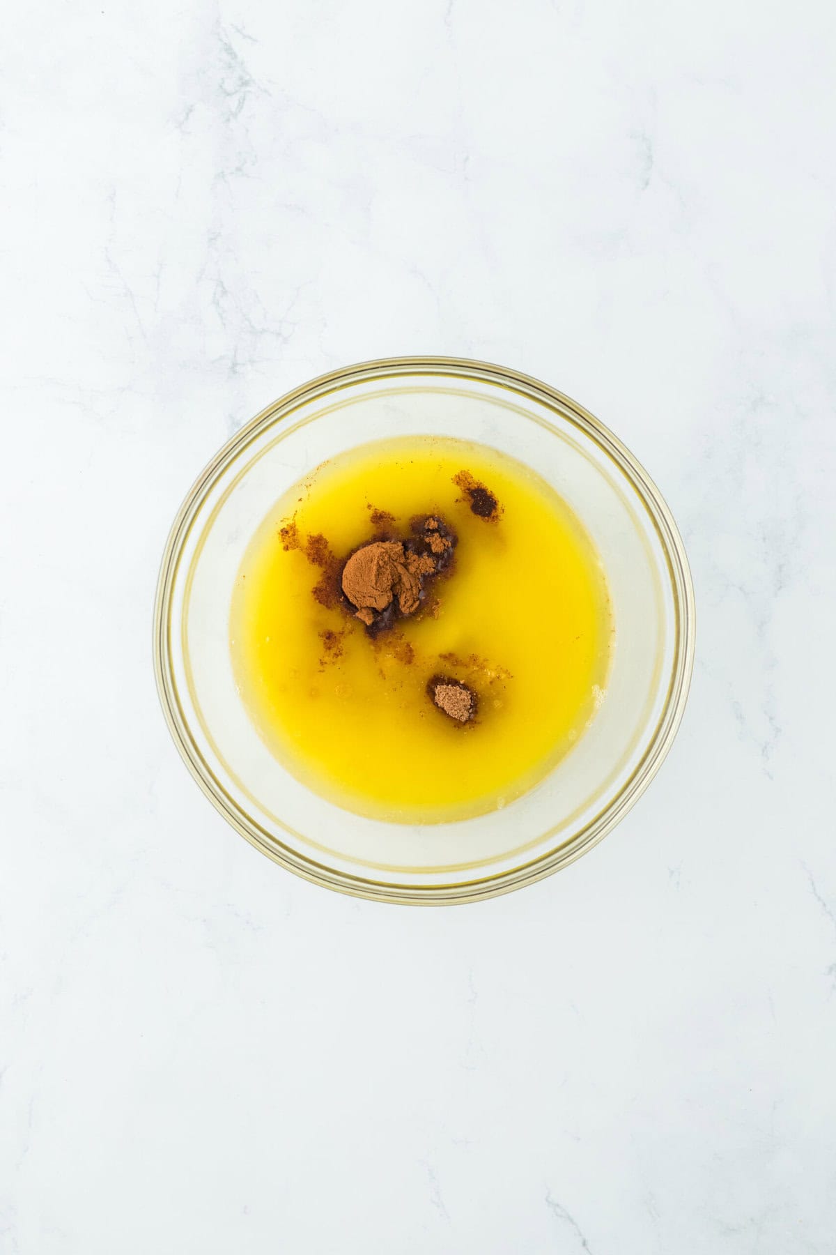 A mixture of melted butter, syrup, garlic, vanilla, cinnamon and nutmeg in a glass bowl on a white countertop