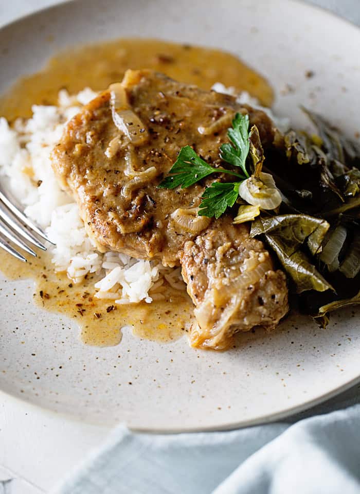 A smothered pork chop swimming in pork chop gravy on white plate with whit rice and greens