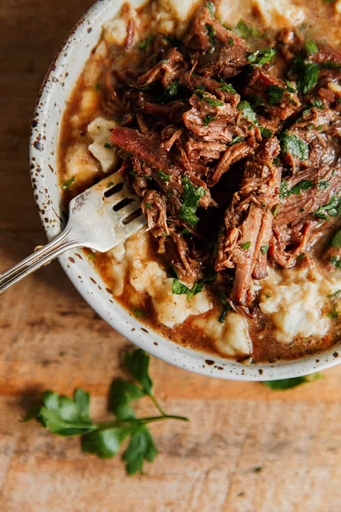 Overhead of a close up of an individual serving of brown sugar Mississippi pot roast in a white bowl with a fork on a bed of mashed potatoes