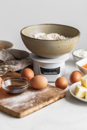 Close up of ingredients for Sweet Potato Cake with Cinnamon Swirl Recipe including eggs on a wooden cutting board, a bowl of flour, butter and a whisk