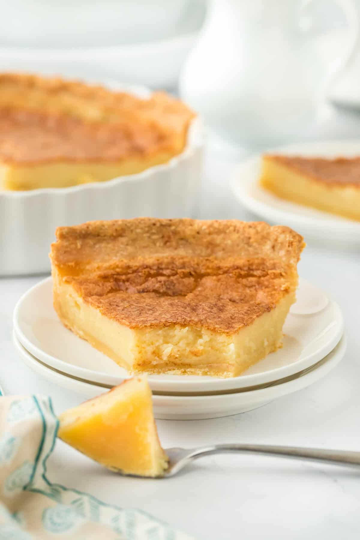 Southern buttermilk pie recipe slice on a white plate with pie in the background and a fork lifting some up