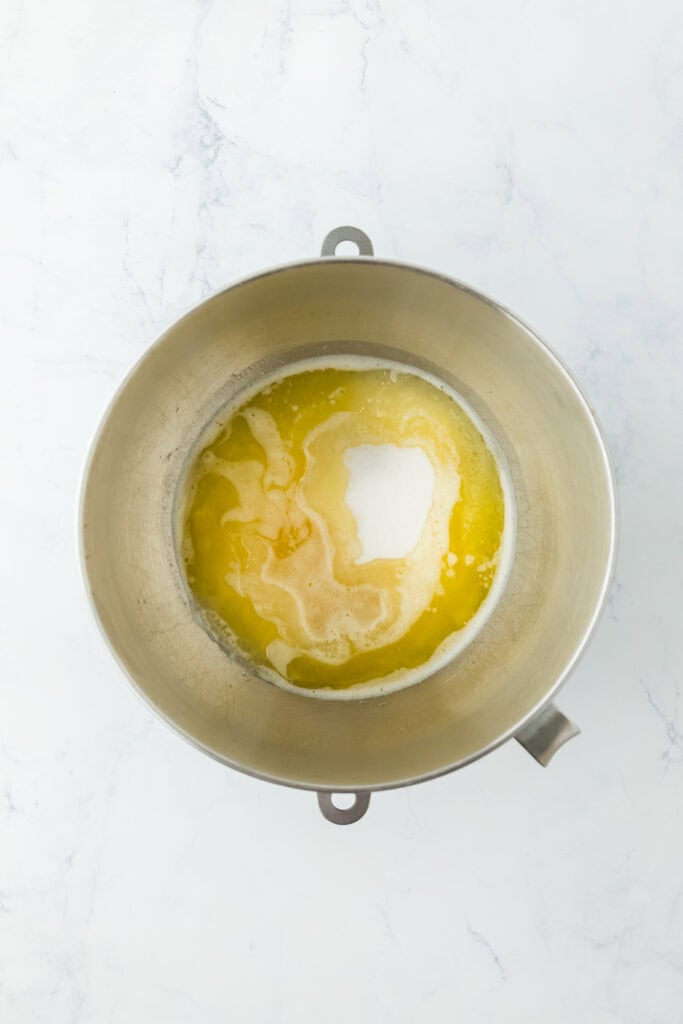 Melted butter and sugar added to a stand mixer bowl on a white countertop