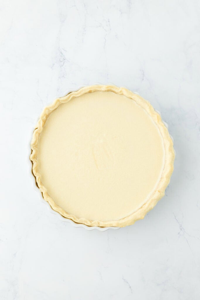 Buttermilk custard filling added to pie crust on white countertop.