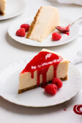 Two Creamy vanilla cheesecake slices on two white plates with raspberry sauce and fresh raspberries