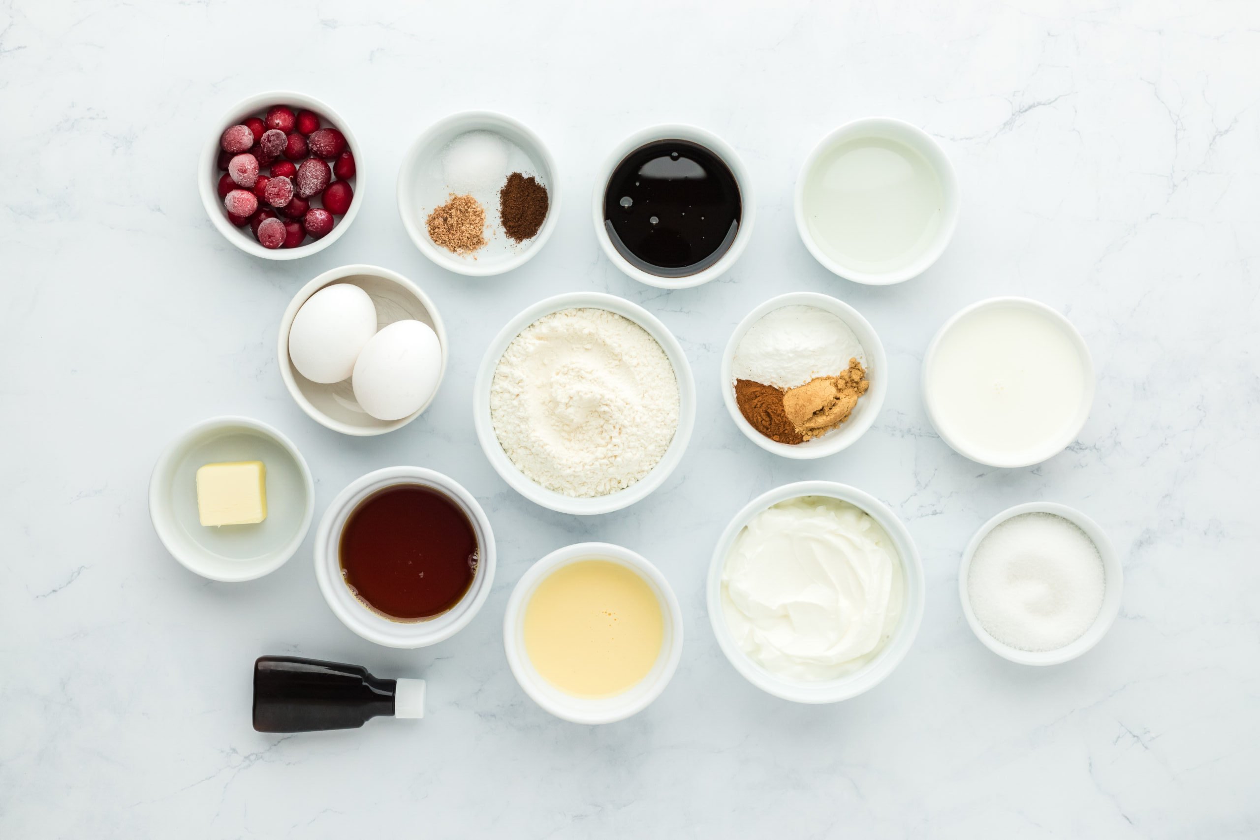 Ginger, molasses, butter, flour, eggs, butter, spices and candied cranberries in white bowls on white countertop