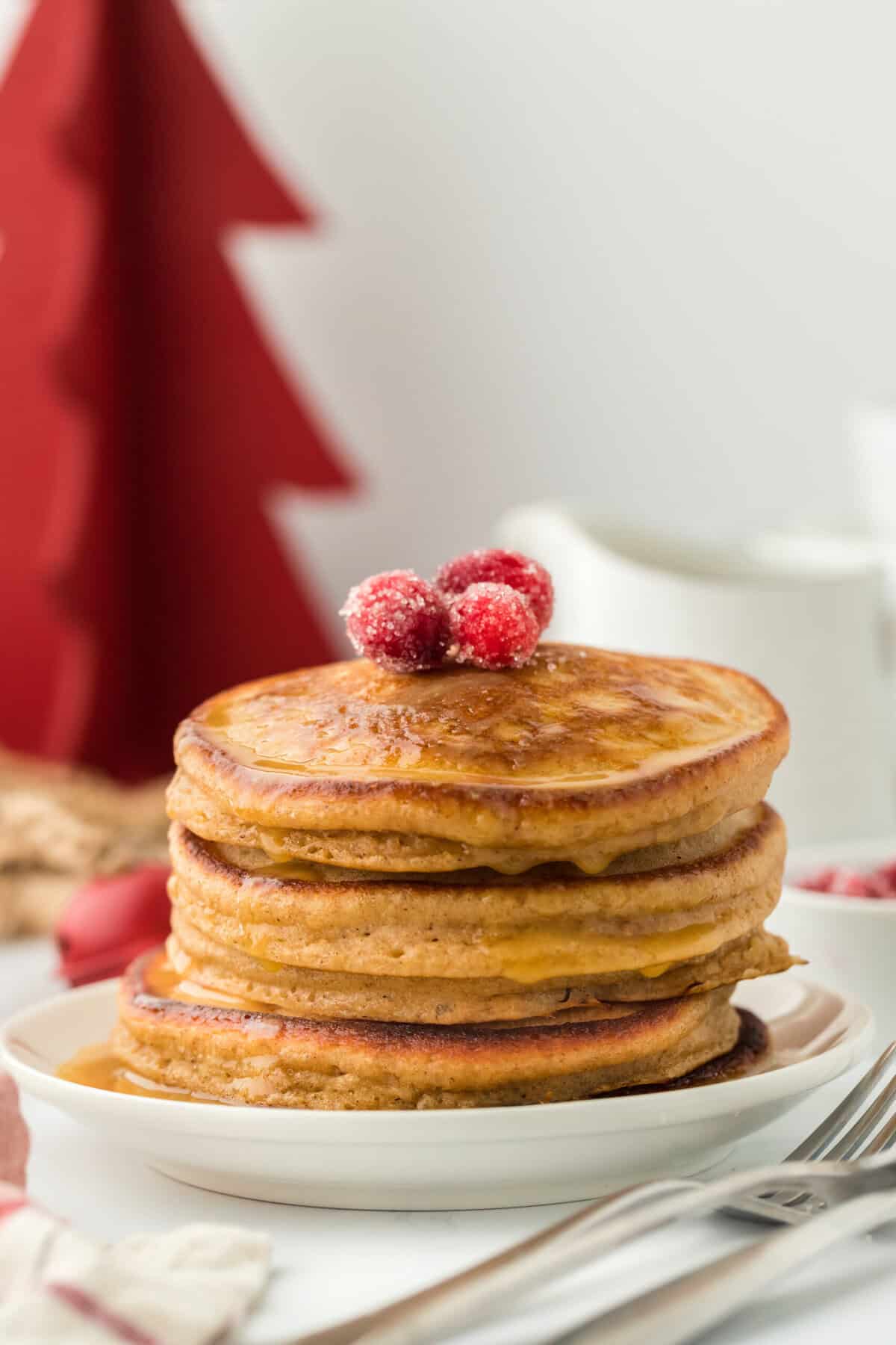 A stack of ginger pancakes with candied cranberries on top in white background with red Christmas tree in the background