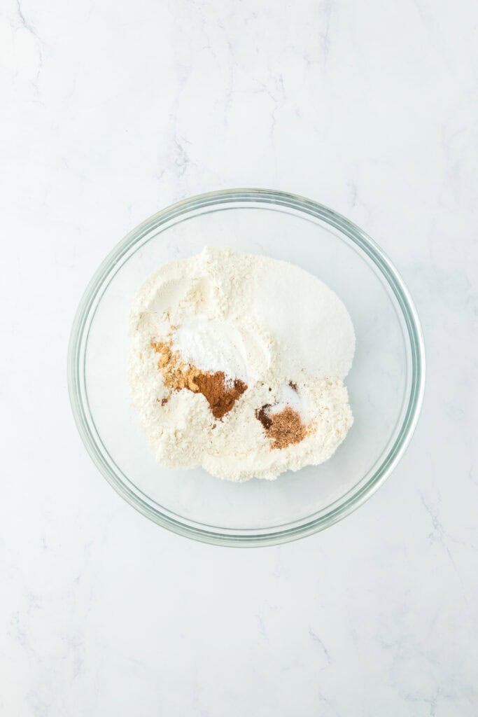 Flour, sugar, baking powder and warm spices in a glass bowl on white countertop