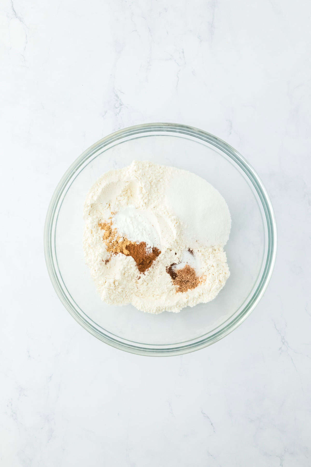 Flour, sugar, baking powder and warm spices in a glass bowl on white countertop