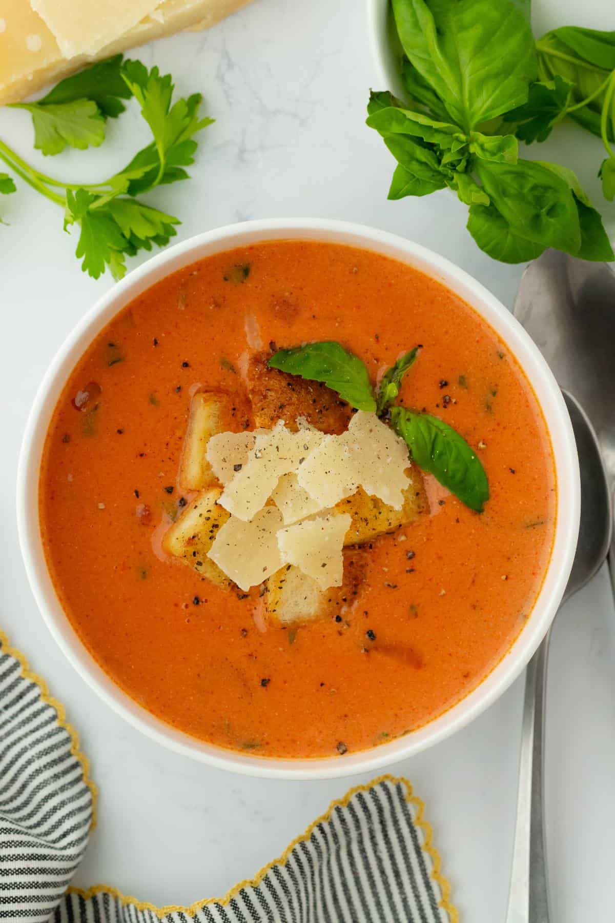 A bowl of homemade tomato basil soup with parmesan, basil, and croutons, styled with fresh herbs and cheese in the background