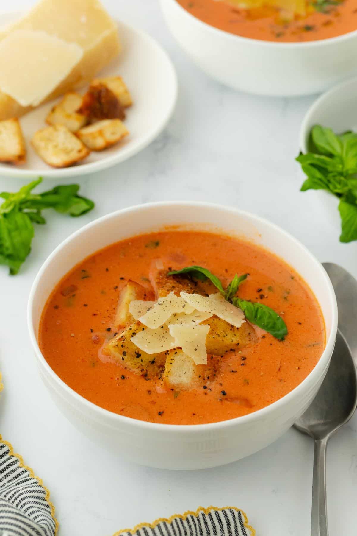 A bowl of homemade tomato basil soup topped with parmesan shavings and basil, served with croutons and more cheese on the side