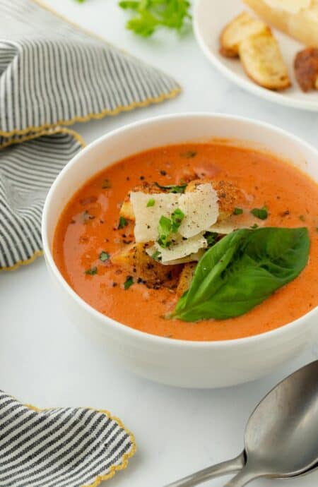 Homemade tomato basil soup garnished with fresh basil, cheese and croutons, presented in a bowl with a spoon beside it