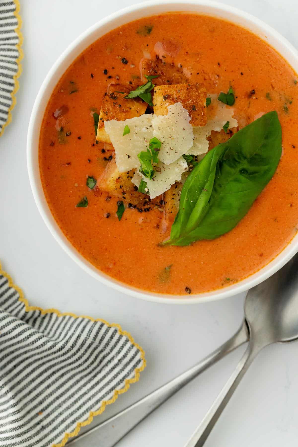 Closeup of the homemade tomato basil soup showing its creamy texture, cheese, basil garnish, and crispy croutons, with spoons and a napkin nearby