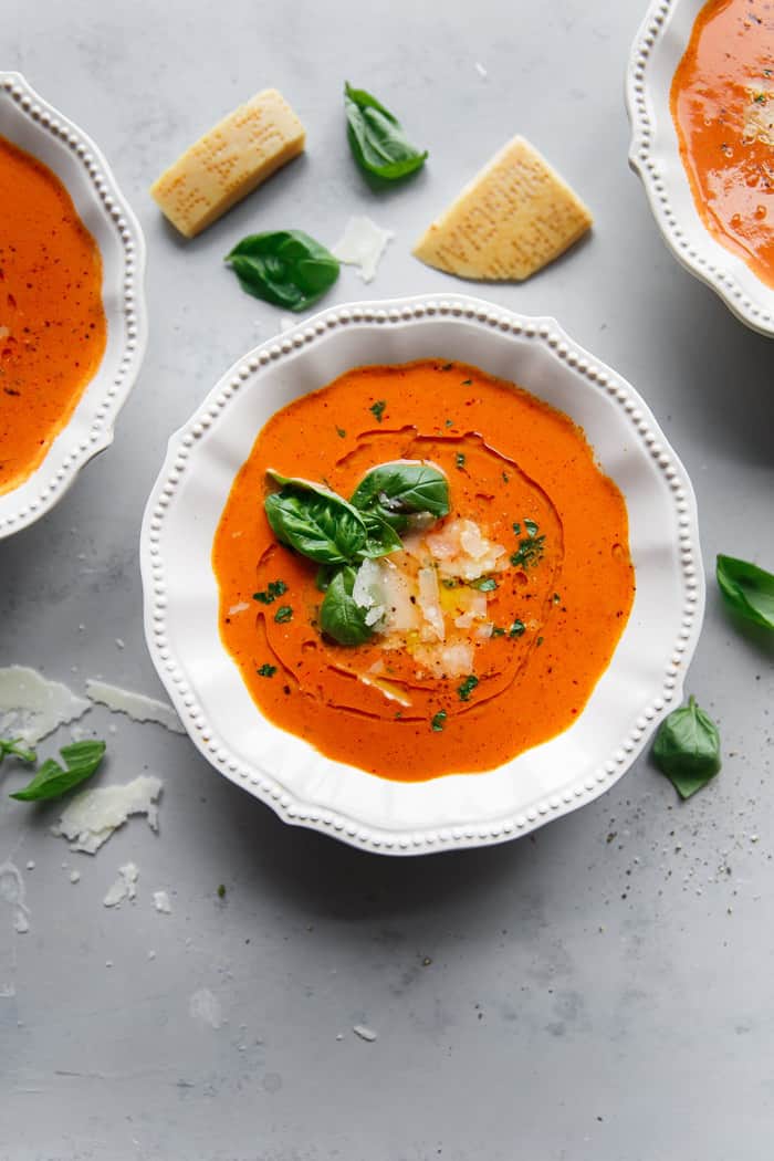 Homemade Tomato Basil Soup recipe in white bowl garnished with fresh basil, oil and shredded parmesan cheese with three bowls of soup