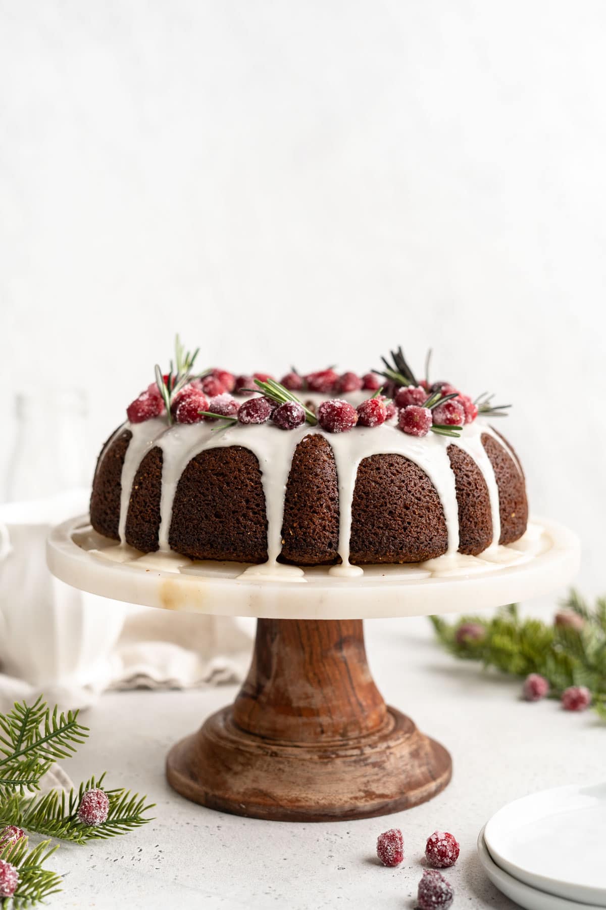 A gingerbread bundt cake on a cake stand garnished with sugared cranberries and rosemary sprigs.