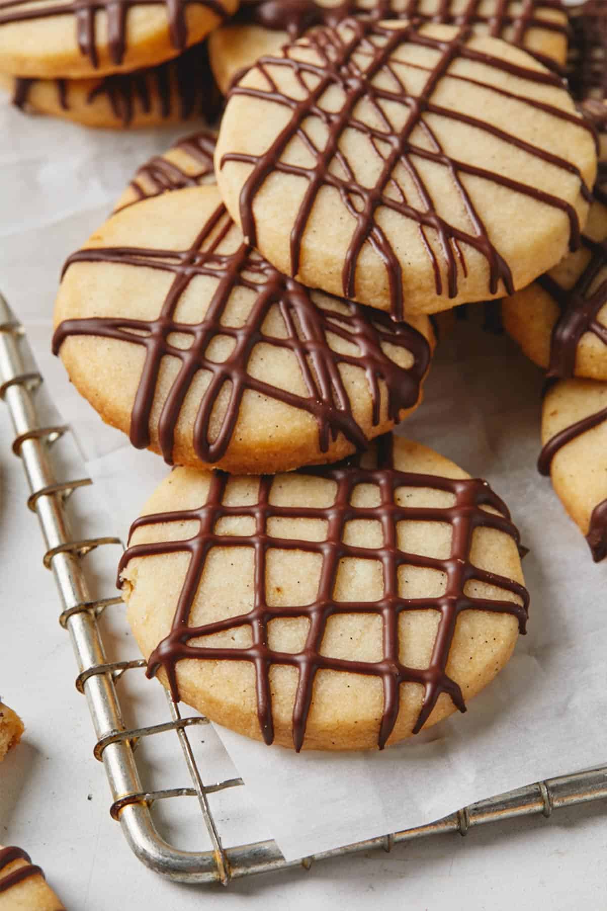 Chocolate drizzled shortbread cookies on a parchment lined wire rack against a white background.