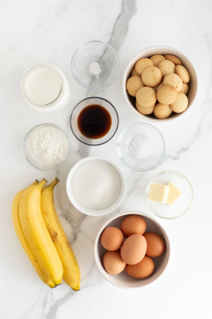 Nilla wafers, bananas, eggs, and ingredients in glass bowls on white countertop