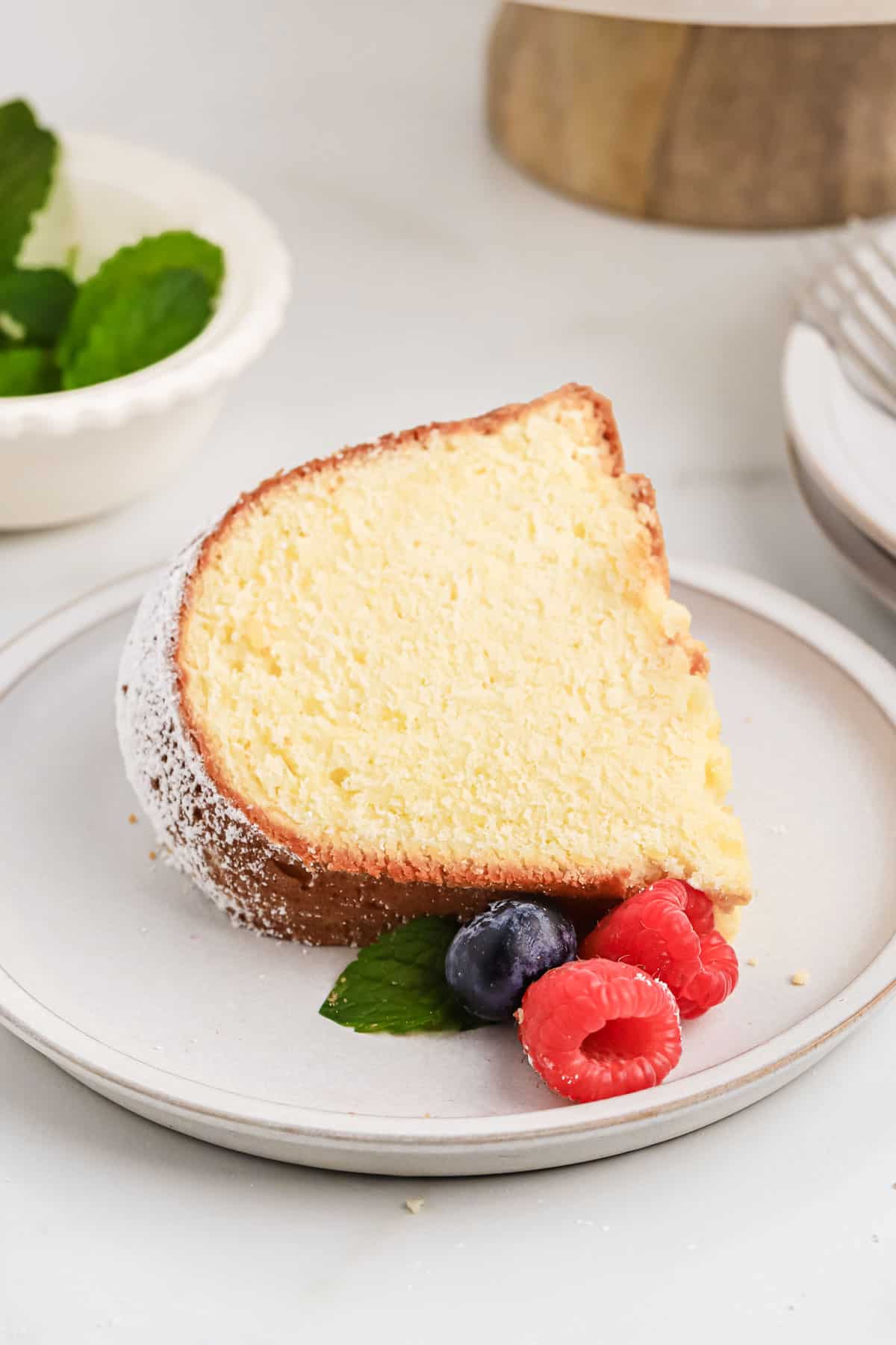 A piece of cream cheese pound cake on a plate with fruit, held by a woman with a fork.