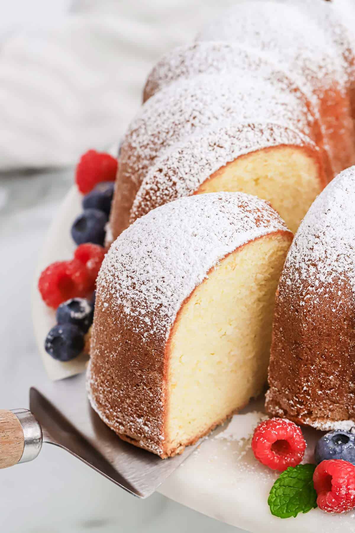 A cream cheese pound cake recipe on a white cake stand with a spatula pulling a slice out surrounded by berries on white background