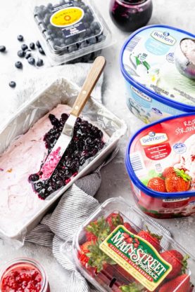 Side shot of blueberry sauce being spread on strawberry ice cream with cartons of ice cream and berries