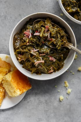 Overhead of a white bowl filled with Greens with hamhock against gray background with cornbread