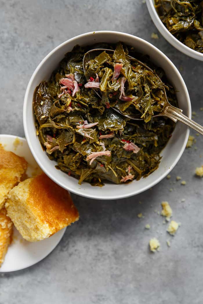 Overhead of a white bowl filled with Southern Collard Greens with hamhock against gray background with cornbread
