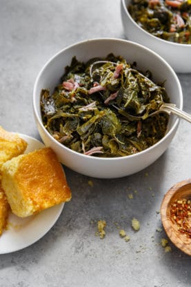 Side shot of a white bowl filled with Collard Greens with hamhock against gray background with cornbread