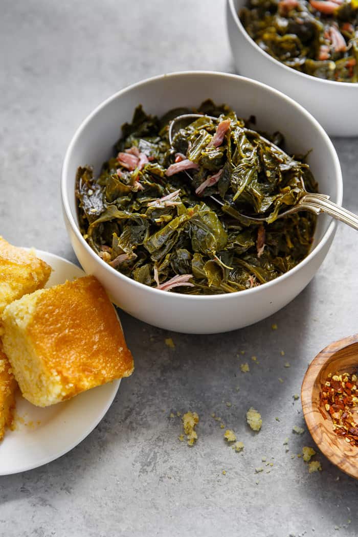 Side shot of a white bowl filled with Southern Collard Greens with hamhock against gray background with cornbread