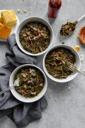 Overhead of three white bowls filled with Southern Collard Greens with hamhock against gray background with cornbread and hot sauce