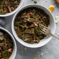 Overhead of three white bowls filled with Southern Collard Greens with hamhock against gray background