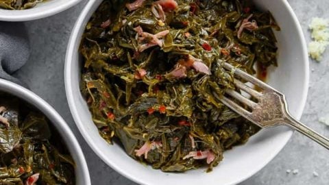 Overhead of three white bowls filled with Southern Collard Greens with hamhock against gray background