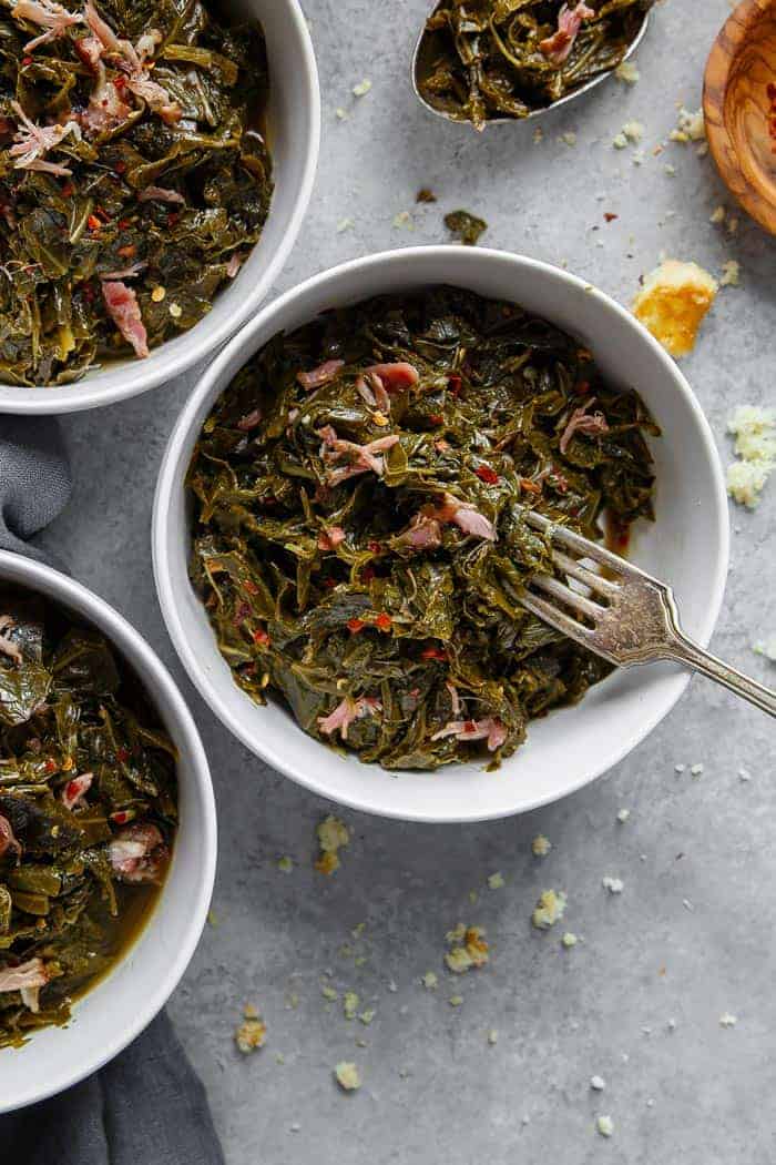 Overhead of three white bowls filled with Southern Collard Greens with ham hock against gray background