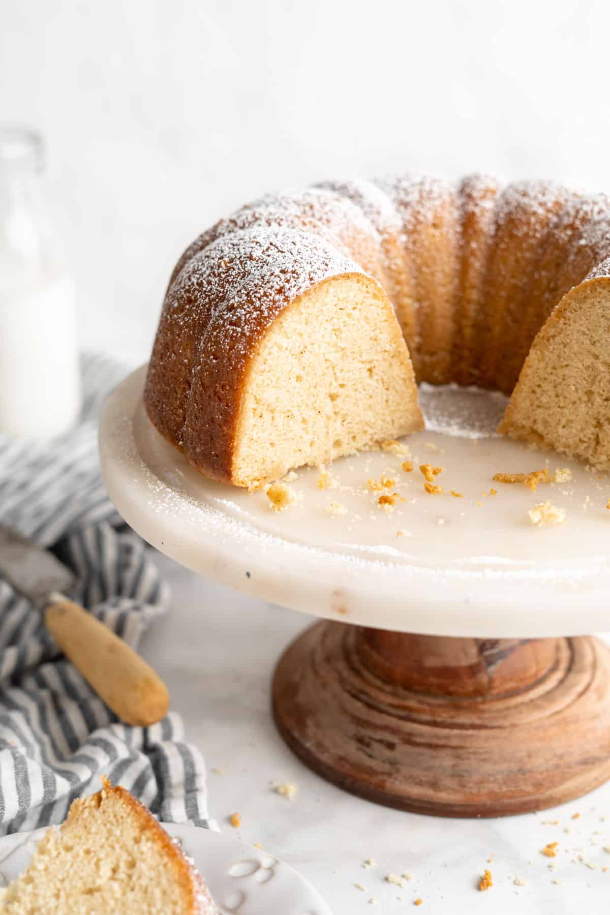 Sliced browned butter cake on cake stand with crumbs and a peak of a slice of cake on a plate.