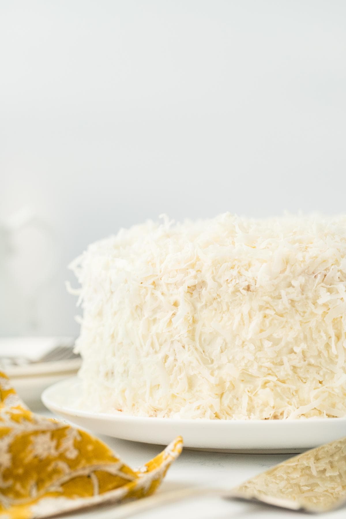 A full, frosted pineapple coconut cake topped with shredded coconut on a white plate with a light background