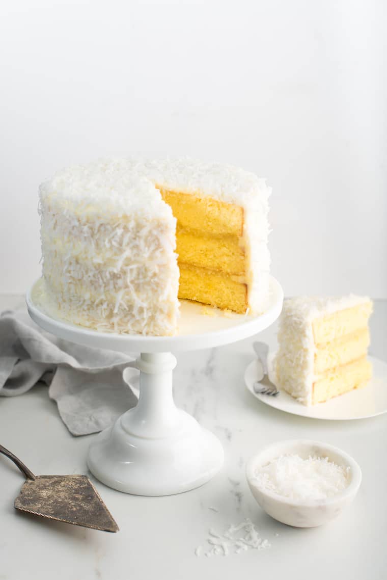 Pineapple Coconut Cake on white cake stand with slice of cake on white plate against white background