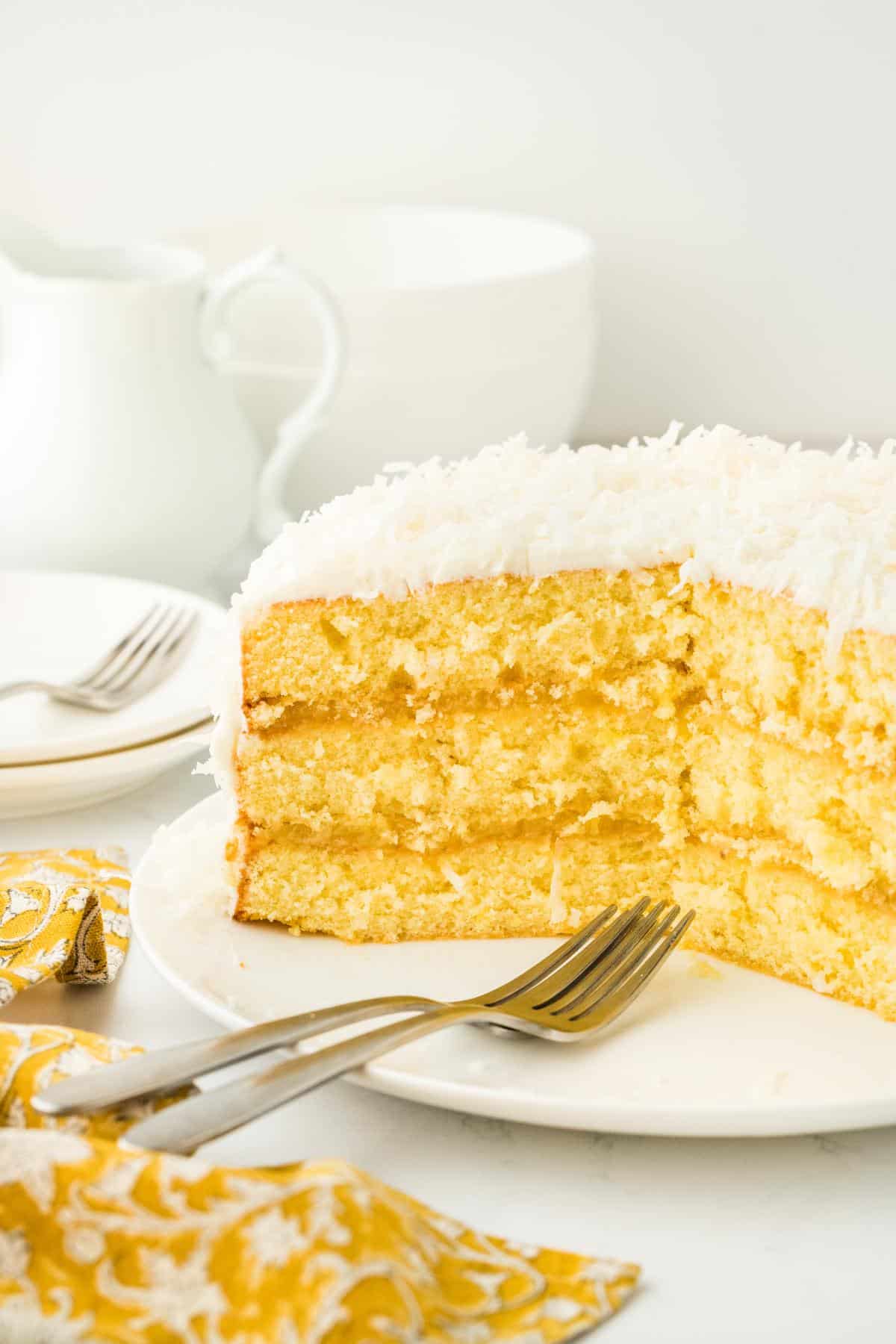 A cross-sectional view of the pineapple coconut cake, showing its moist yellow layers and creamy frosting, with forks next to it and more plates and forks in the background