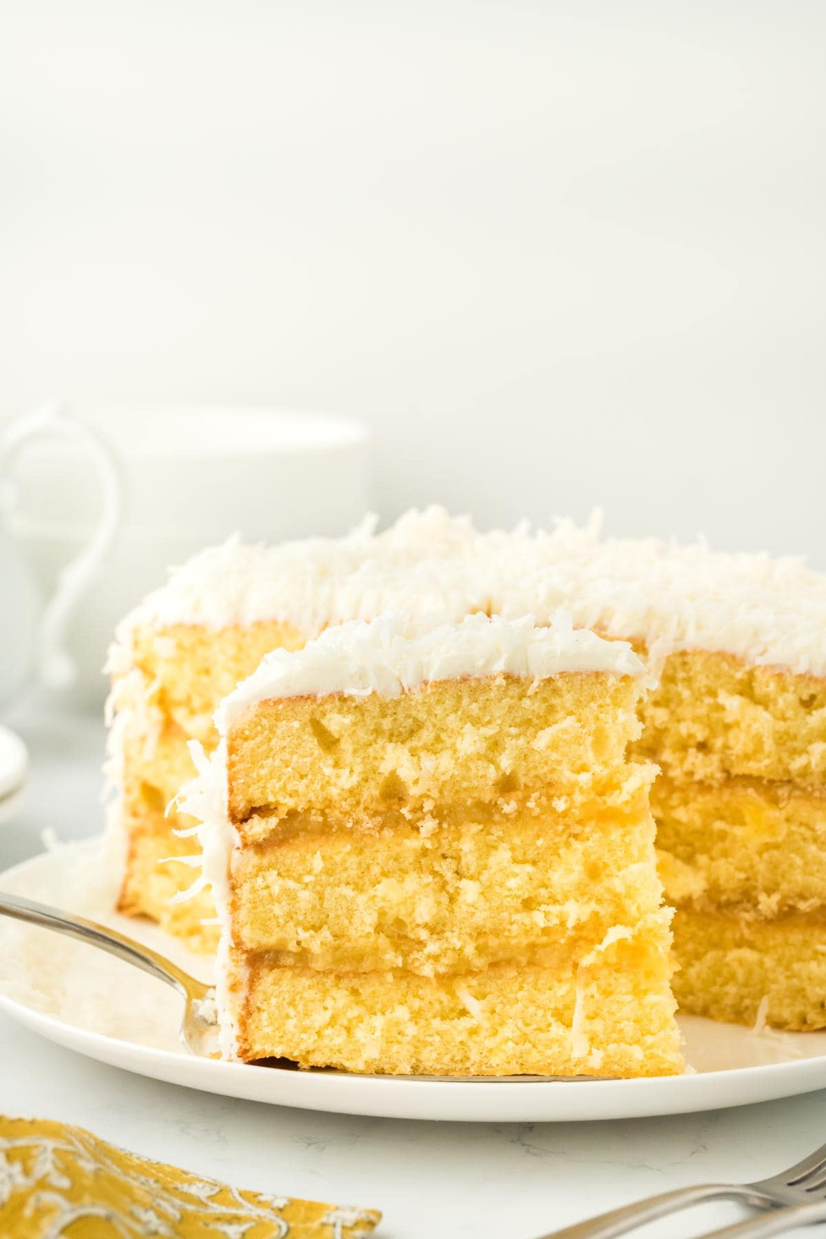 A slice of pineapple coconut cake being served with a fork, with the rest of the cake behind it