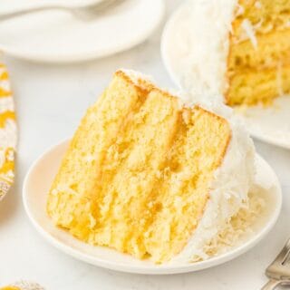 A closeup of a single slice of pineapple coconut cake on a plate, emphasizing the texture of the layers and frosting, with plates and the rest of the cake in the background