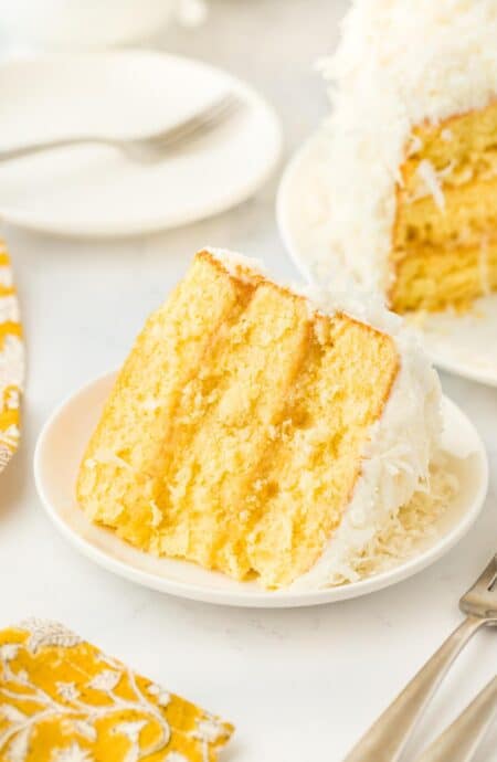 A closeup of a single slice of pineapple coconut cake on a plate, emphasizing the texture of the layers and frosting, with plates and the rest of the cake in the background