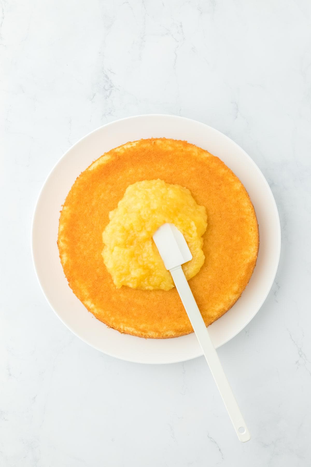 A cake layer on a plate with pineapple filling being spread using a spatula